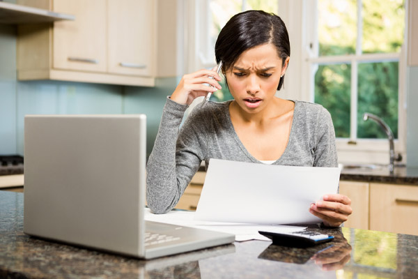 A concerned woman sits at a kitchen counter with a laptop and papers.