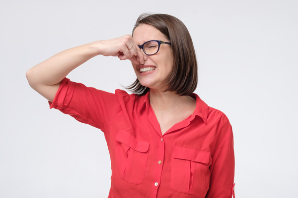 A woman wearing glasses and a red shirt grimaces, holding her nose. 