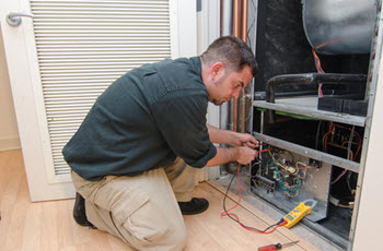 A technician kneels and uses a multimeter to inspect the wiring within an HVAC unit. 