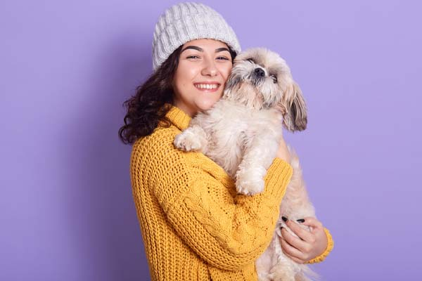 A young woman in a beanie and yellow sweater holds a small dog against a solid purple background. 