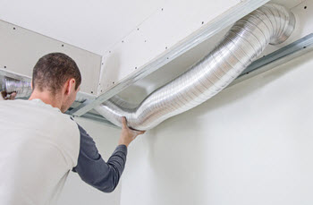 A man installs a flexible metal duct into a ceiling framing.