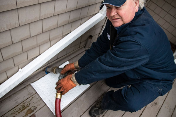 A man wearing work clothes connects a hose to an outdoor tank; an absorbent mat rests beneath. 