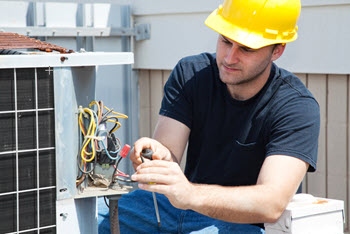 A technician wearing a hard hat repairs an outdoor air conditioning unit. 