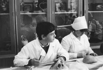 Two students in lab coats are working at a table, with anatomical models displayed behind them.