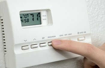 A close up shows a hand pressing the "PROG/MAN" button on a white thermostat.