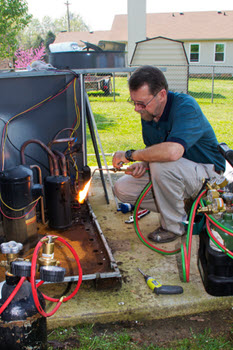 A man in a blue shirt and khaki pants uses a torch to work on a heating or cooling unit outdoors. 