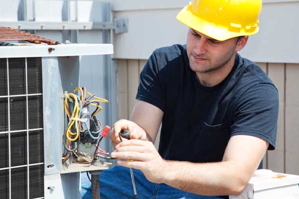 A man wearing a yellow hard hat works on the exposed wires of an outdoor AC unit.