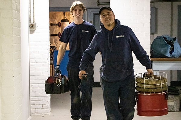 Two HVAC technicians wearing company branded shirts carry toolbags towards the camera.