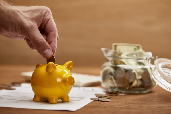 man dropping coin in a small golden piggy bank with bills and coins in the background depicting savings